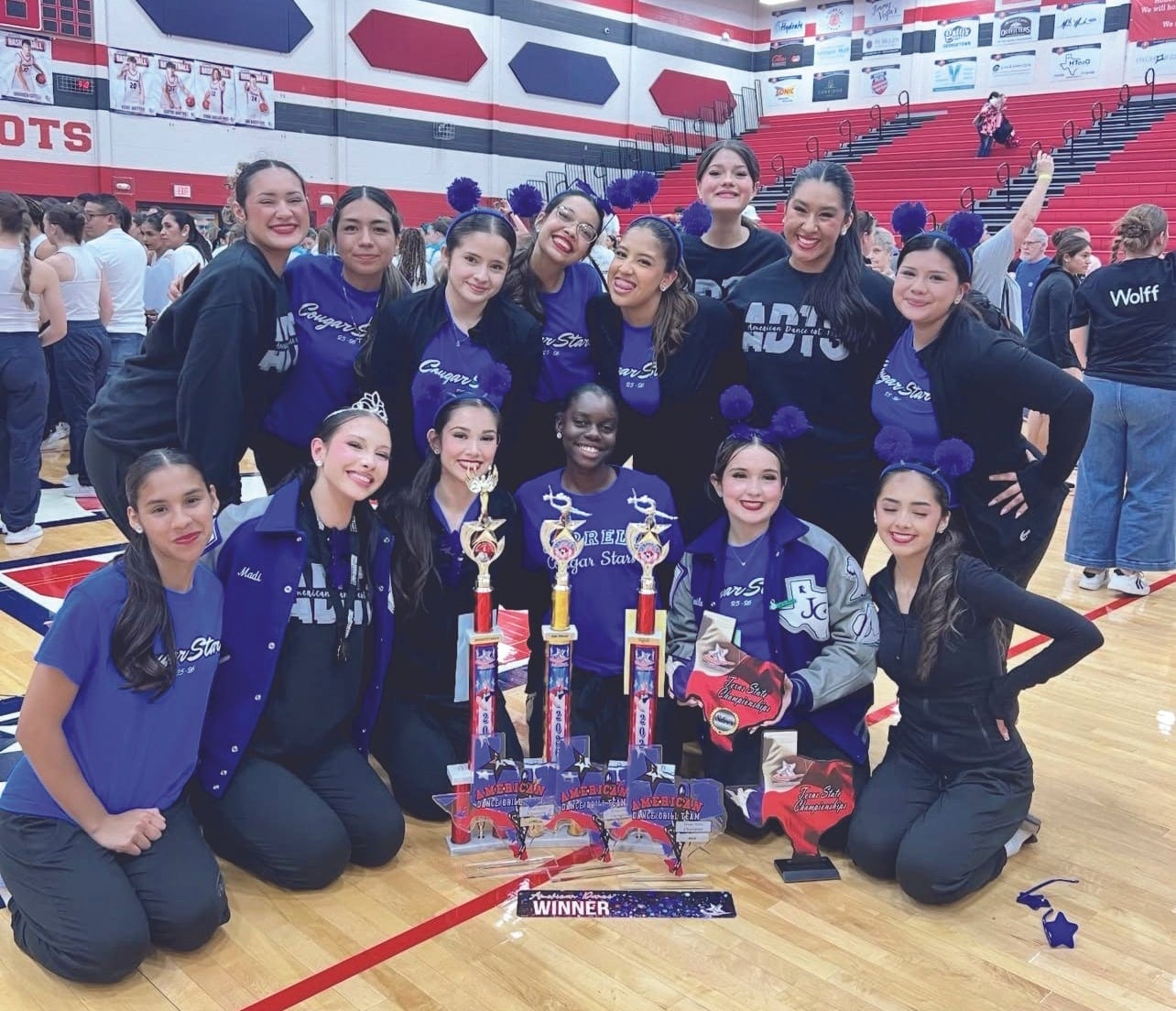 Dance team members in purple and black kneel and stand together in a gym, posing with several tall trophies and a winner banner.