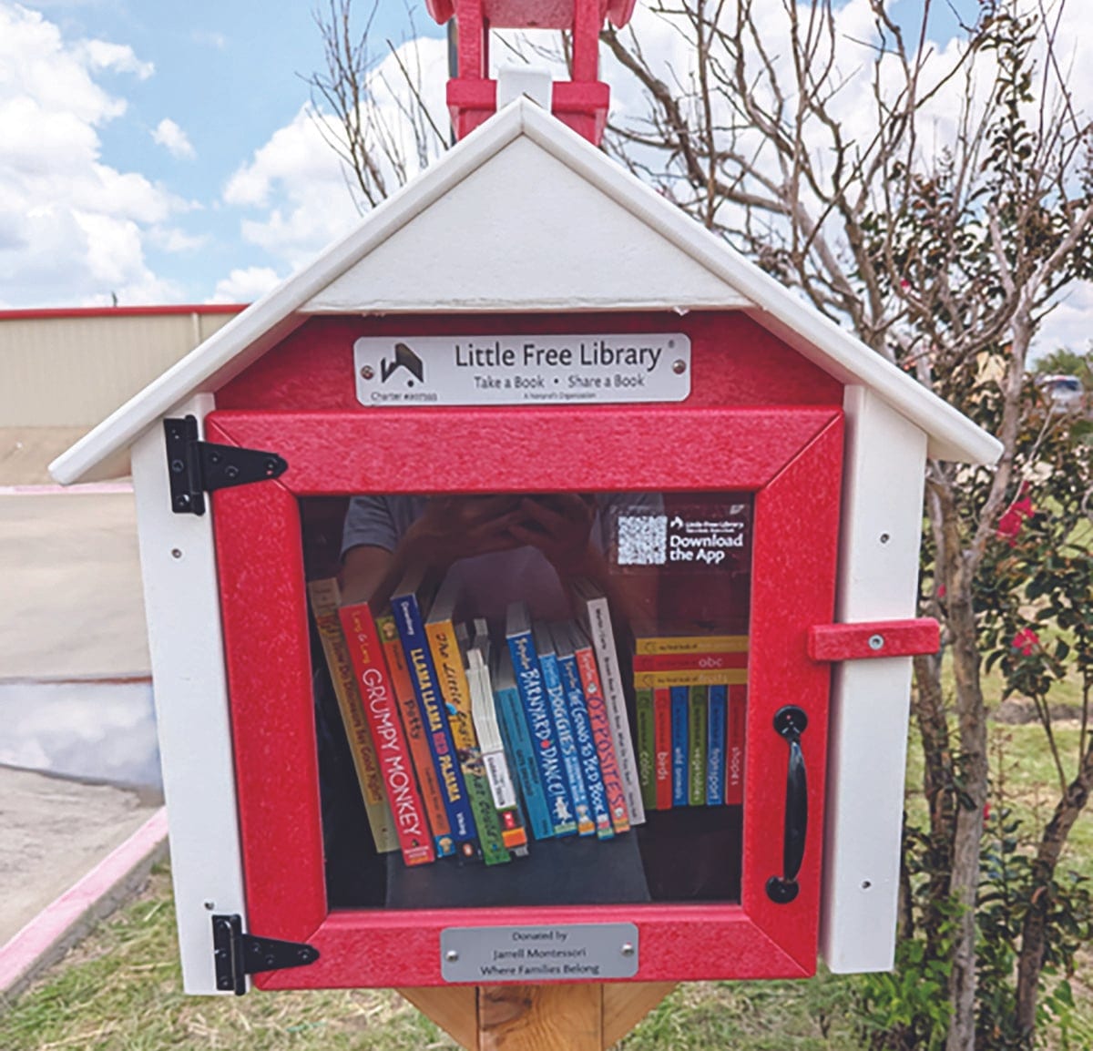 Close-up of a red and white schoolhouse-shaped Little Free Library full of children's books, with a plaque reading Donated by Jarrell Montessori.
