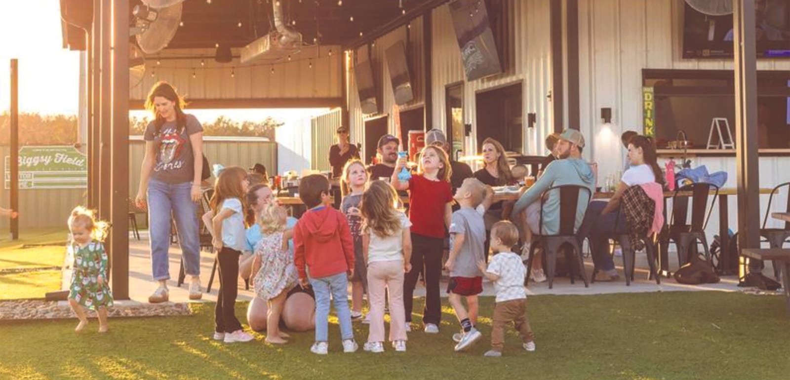 A group of young children gathers around a woman on artificial turf at an open-air restaurant patio while adults sit at tables in the background at sunset.