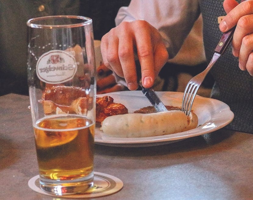 A glass of beer on a coaster beside a plate where someone cuts into a white sausage with a knife and fork.