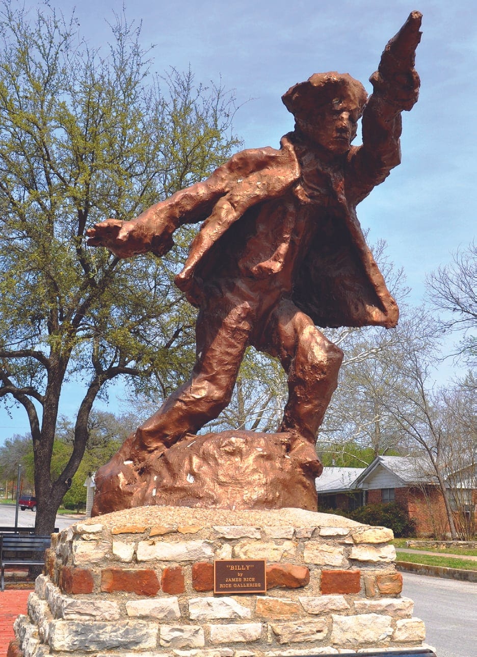 A copper-colored statue of Billy the Kid with arm raised, standing on a stone and brick pedestal along a tree-lined street