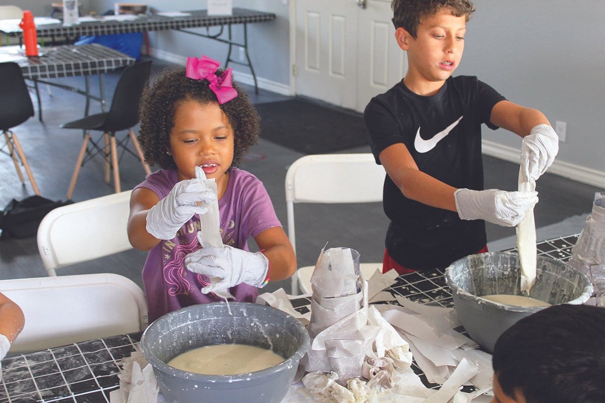 Two children in white gloves dip strips of paper into bowls of paste during an art project at a table.