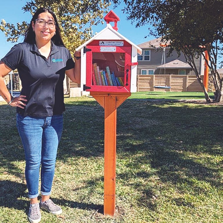 A smiling woman in glasses and a dark polo stands beside a red and white Little Free Library on an orange post in a grassy park.