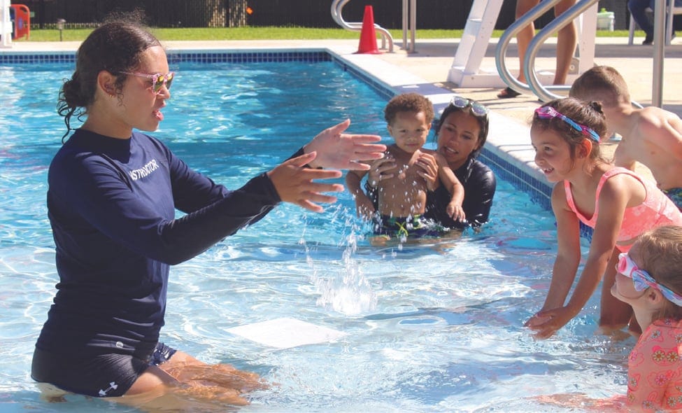 A swim instructor in a navy rash guard leads young children through a lesson in a sunny pool.