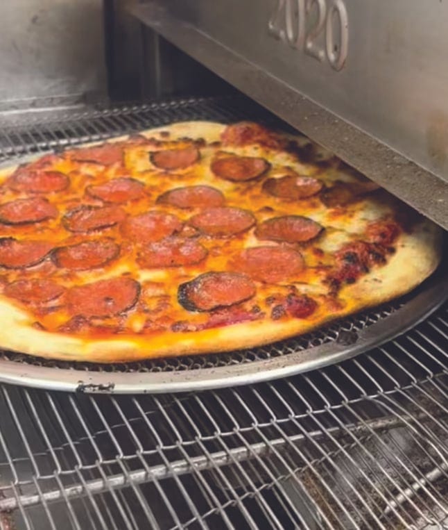 A pepperoni pizza with a golden crust coming out of a conveyor pizza oven.