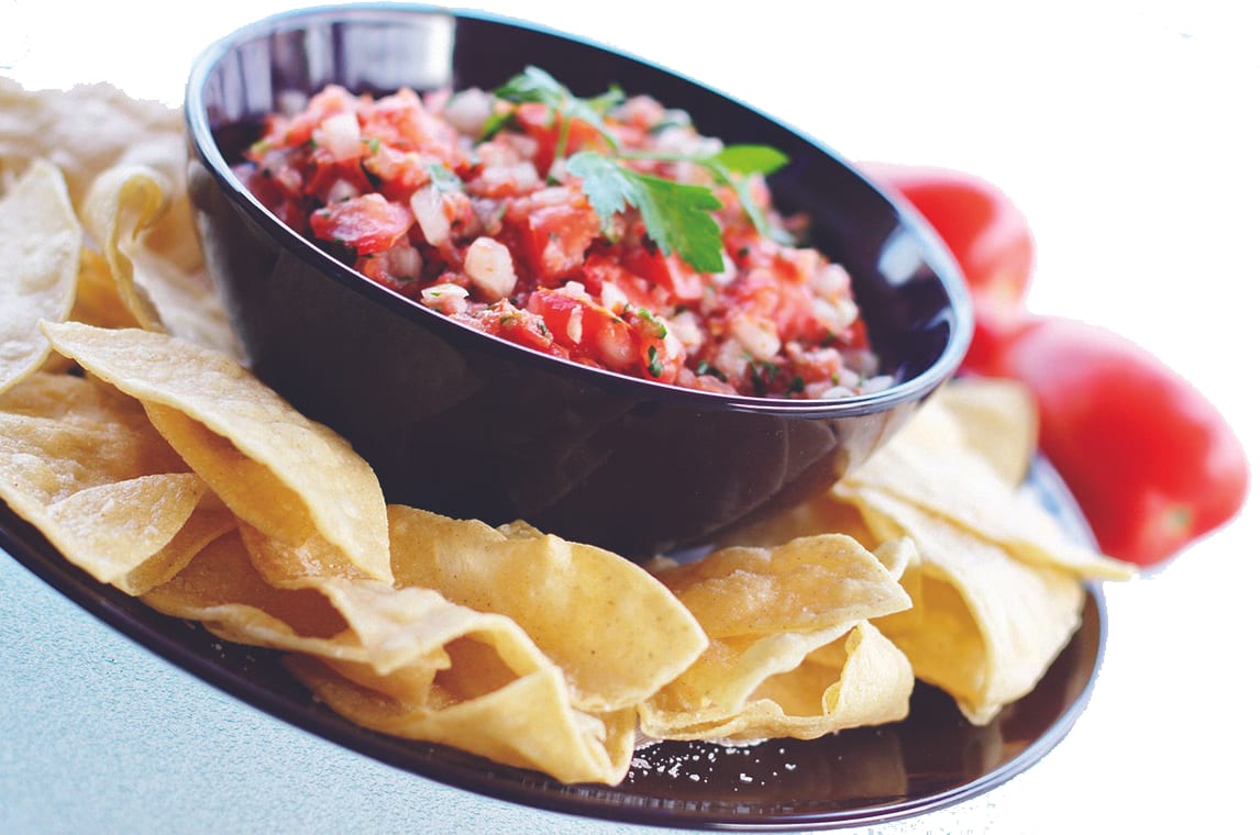 A black bowl of fresh chunky salsa topped with cilantro, surrounded by tortilla chips with tomatoes in the background.
