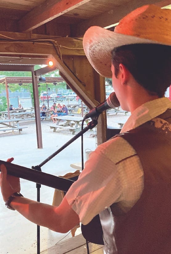 A performer in a straw cowboy hat and vest sings into a microphone under a wooden pavilion, with picnic tables and guests in the background.