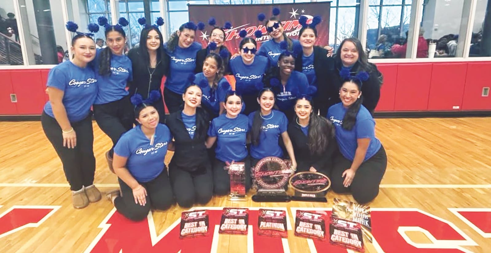 The Cougar Stars in blue team shirts and blue pom-pom headbands pose on a gym floor with awards, including several Best in Category banners.