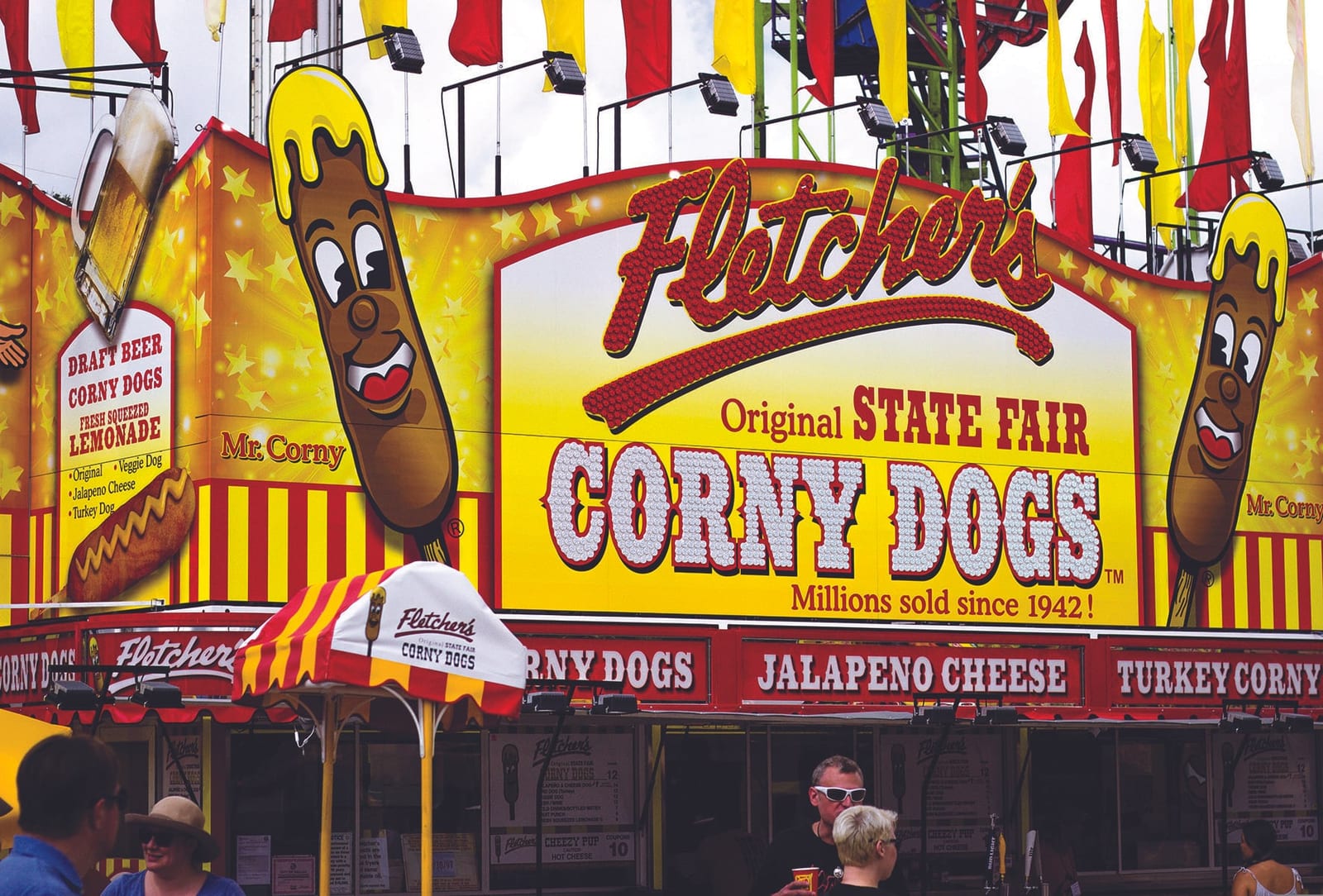 Brightly lit Fletcher's Original State Fair Corny Dogs stand with cartoon corny dog mascots and fairgoers in front