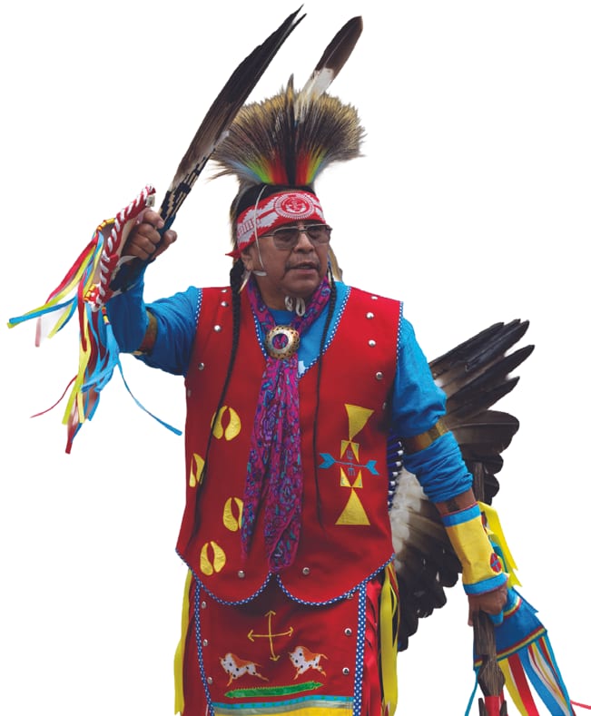 A Native American dancer in a feathered headdress, red beaded vest and blue shirt raises a feathered fan and ribbons mid-dance.