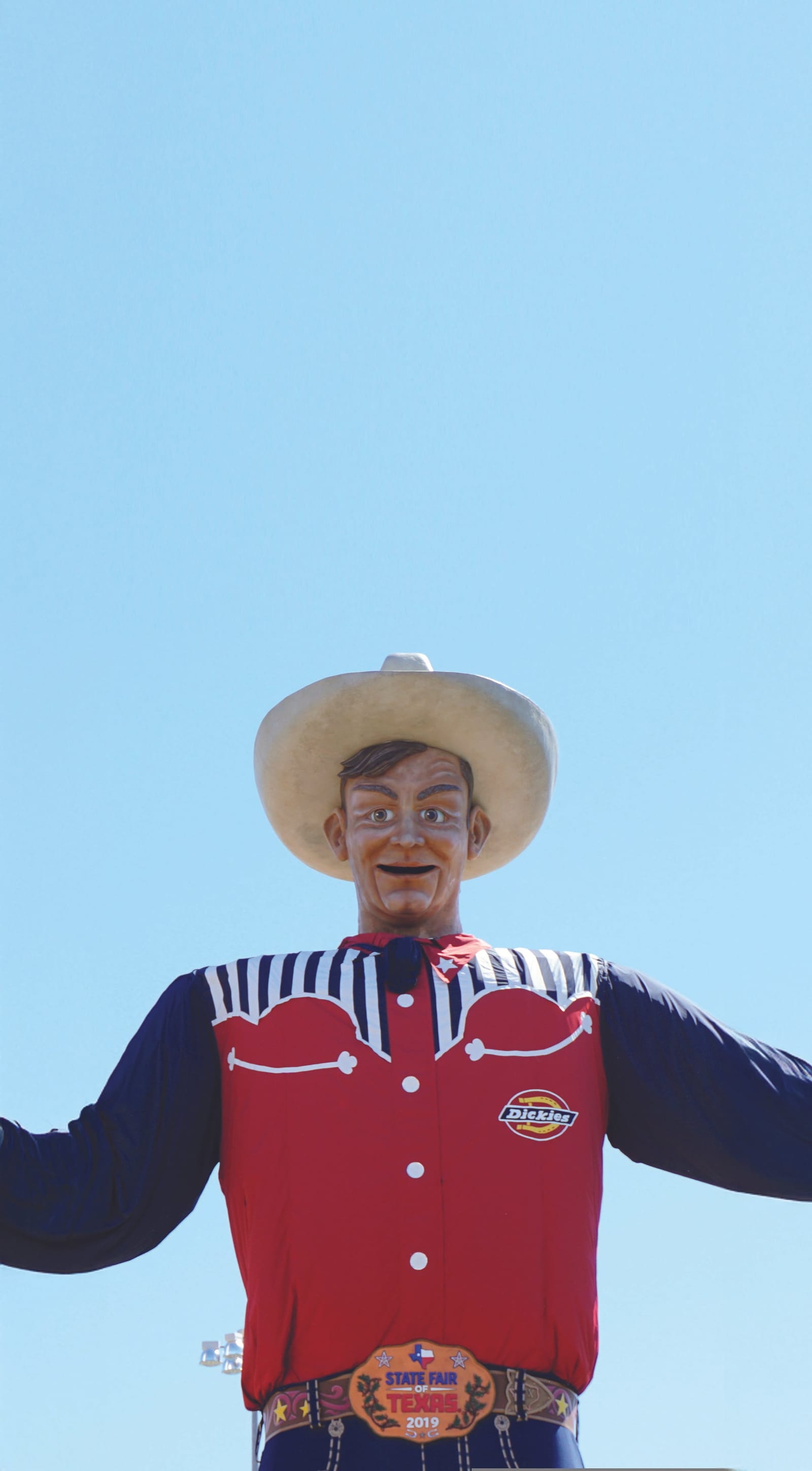 The giant Big Tex cowboy figure in a white hat, red and navy western shirt and State Fair of Texas belt buckle, arms outstretched against a clear blue sky