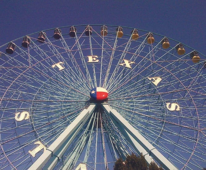 The Texas Star Ferris wheel with large letters spelling Texas and a Texas flag hub against a deep blue sky