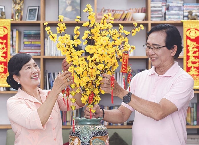 A smiling couple decorates a vase of yellow blossoms with small red ornaments in a room lined with bookshelves and red banners.