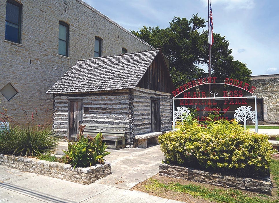 A small historic log cabin beside a stone building, next to a Peppermint Square archway sign, flowering shrubs and a flagpole