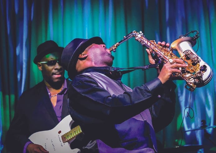 A musician in a hat plays a saxophone on stage against blue and green curtains, with a guitarist in sunglasses behind him.